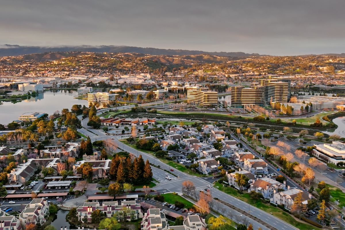 Aerial view of a cityscape with residential areas, winding roads, and office buildings near a water body. Hills and cloudy sky in the background.