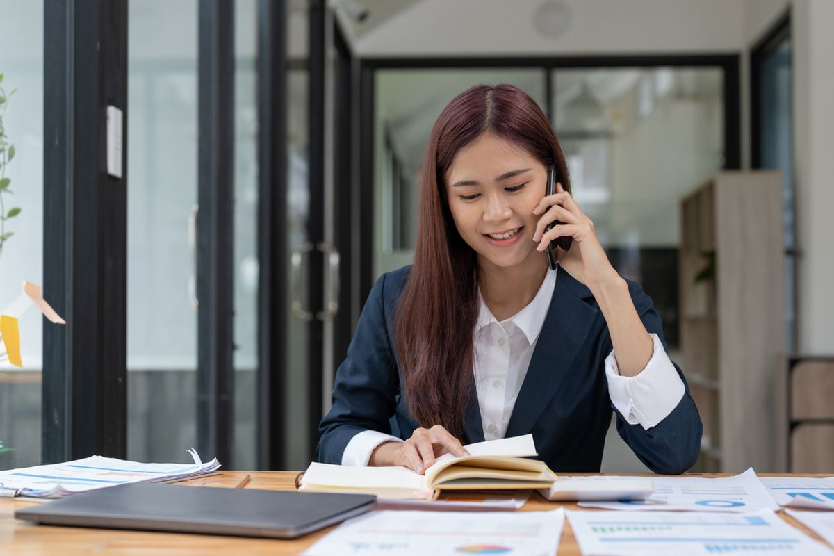 Woman in a suit talks on the phone while reading a book at a desk in a modern office. She is smiling, surrounded by documents.