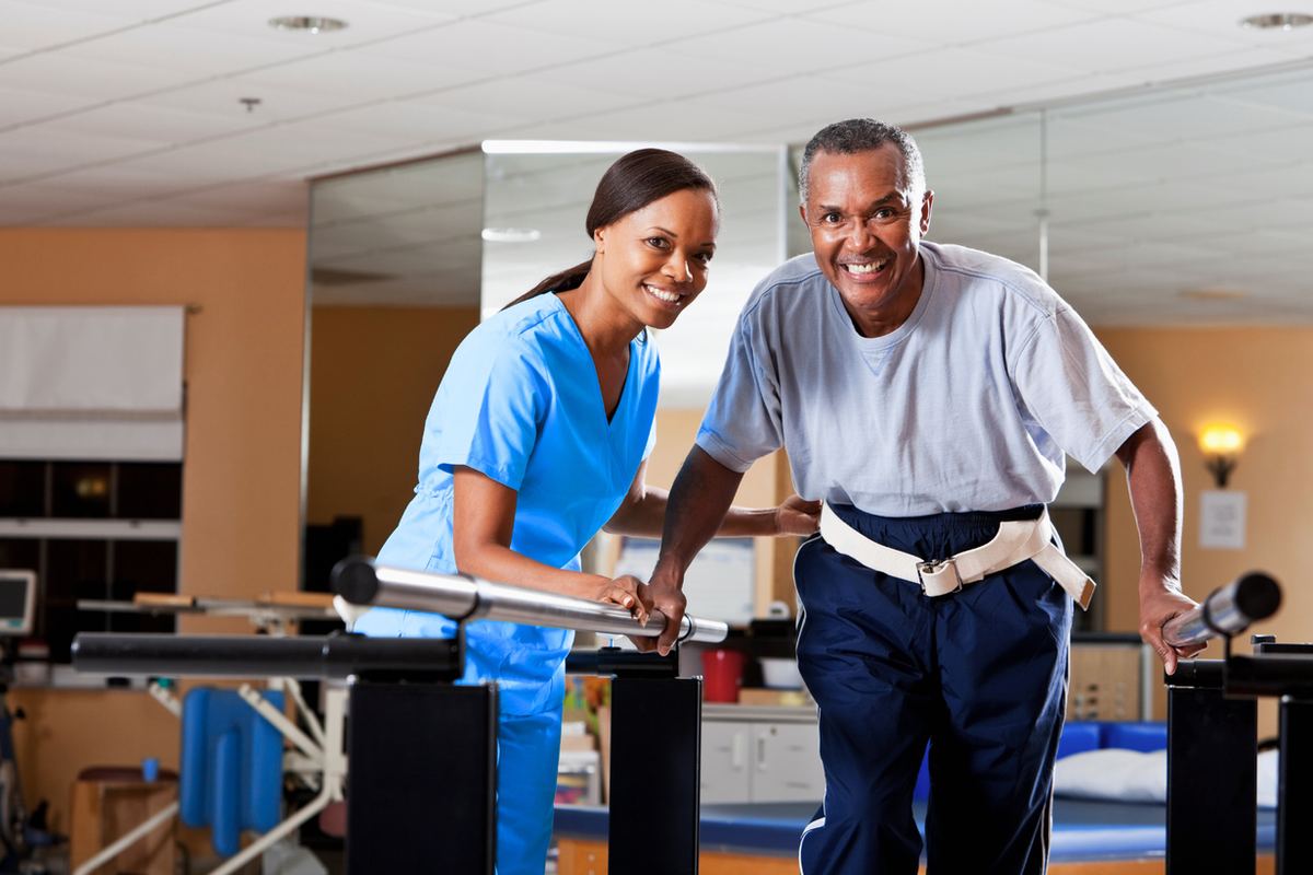 A man in a white shirt uses parallel bars to walk with assistance from a woman in blue scrubs, both smiling in a bright rehabilitation room.