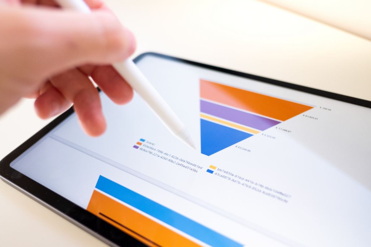 Hand using a stylus to point at a colorful pie chart on a tablet screen. The background is white, creating a bright, analytical setting.