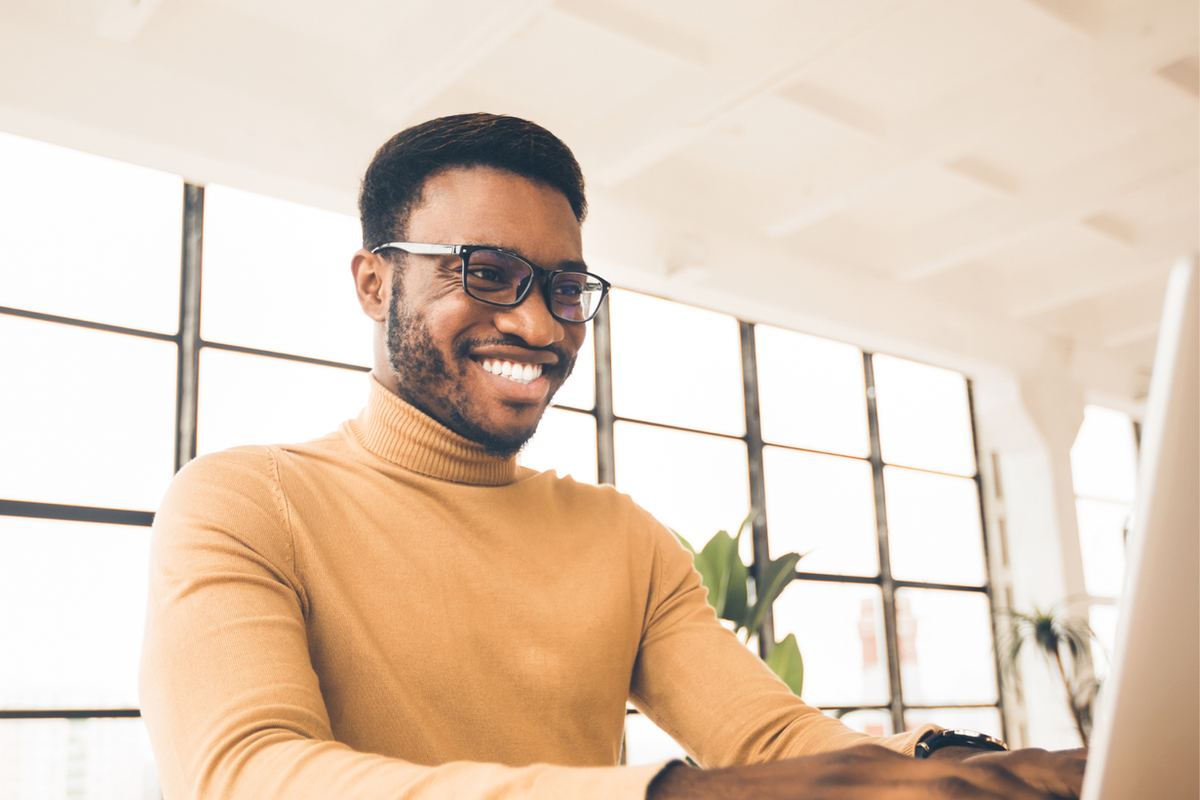 Man in glasses and tan turtleneck smiles while working on a laptop. Bright room with large windows and plants. Relaxed mood.