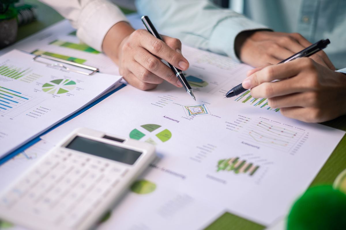 Two people analyze financial charts and graphs on papers. A white calculator lies nearby on a green table, with pens pointing at data.